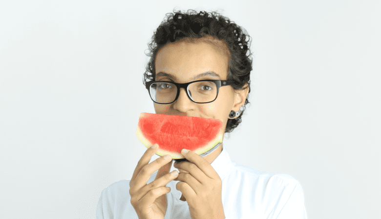 jovem mulher segurando uma fatia de melancia na altura da boca. Ela usa óculos de grau e está vestindo uma blusa branca