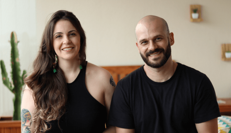 casal sentado pousando para a foto. Ambos vestem camiseta preta e ao fundo há um vaso com um cacto