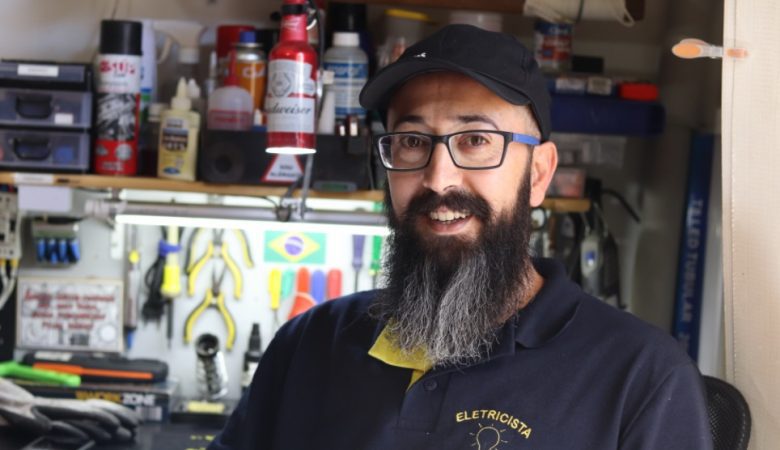 homem sentado em uma cadeira vestindo uma camisa polo preta com detalhes amarelos. Ele também usa óculos e boné preto, e tem barba longa
