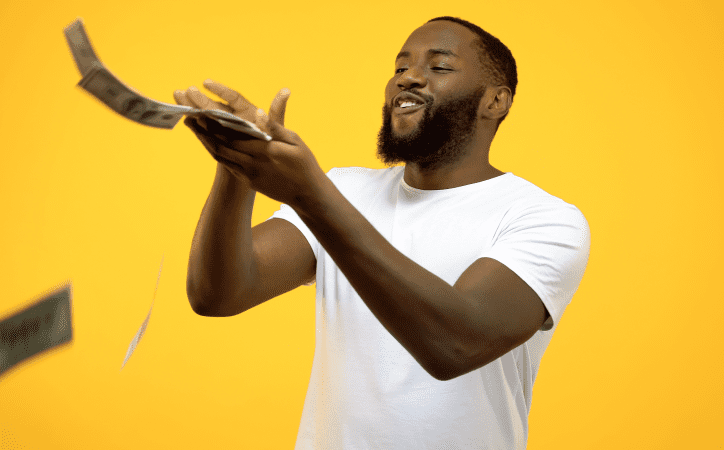 Homem negro vestido de branco jogando notas de dinheiro ao ar com o fundo amarelo.
