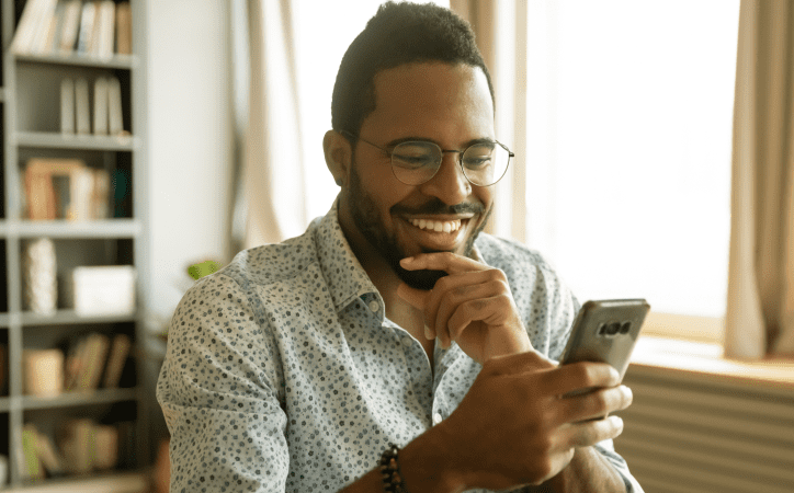 Homem de óculos e barba sorrindo ao mexer em um smartphone. 