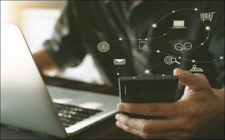 Homem vestido de preto segurando um celular e mexendo em um notebook, com símbolos digitais saindo da tela para ilustração. 