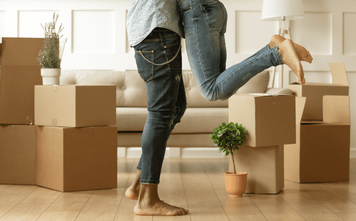 Homem levantando uma mulher, ambos vestidos de jeans, com caixas de mudança, sofá e plantas ao fundo. 