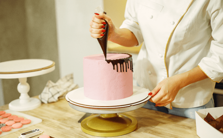 Pessoa decorando um bolo rosa e preto com saco de confeiteiro, em cima de uma mesa com diversos docinhos e utensílios. 