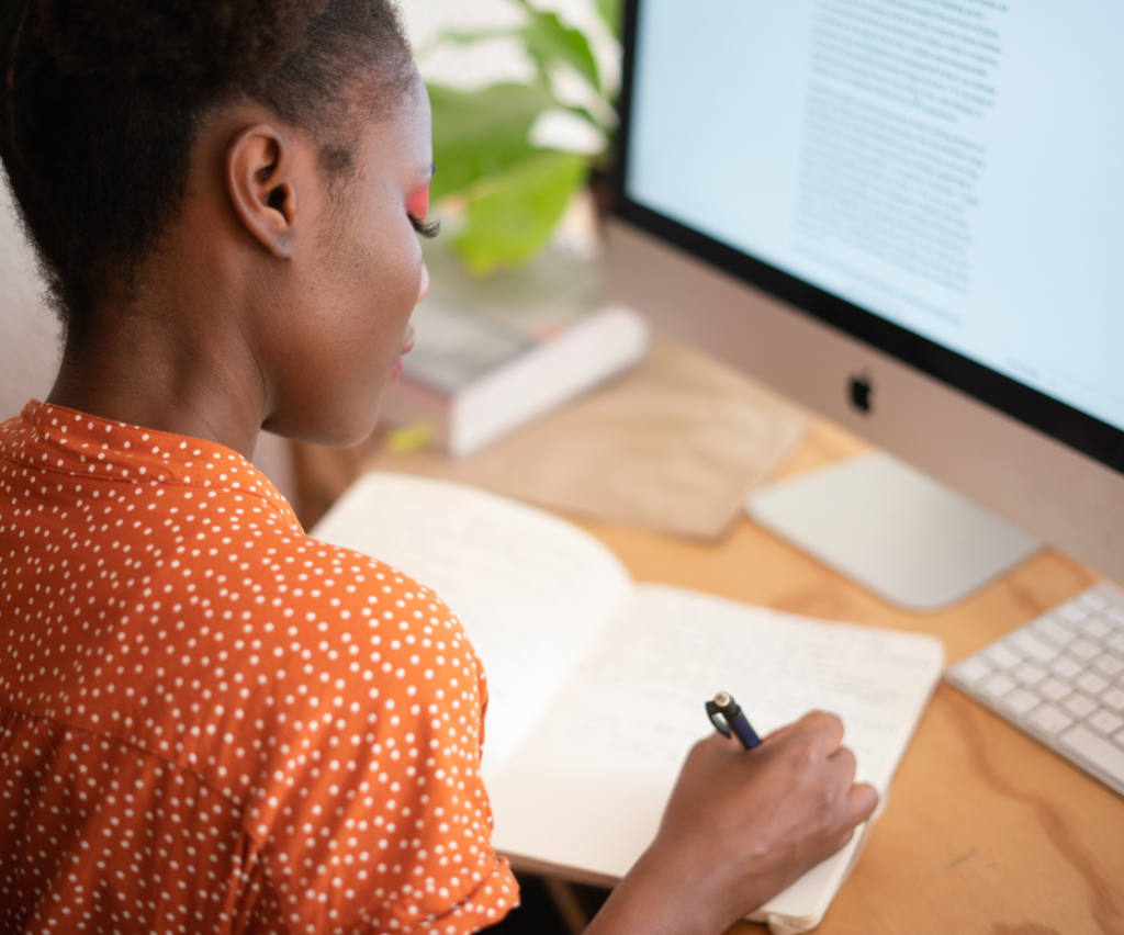 mulher negra vestindo uma camisa laranja. Ela está escrevendo em um caderno e a sua frete a uma tela de computador